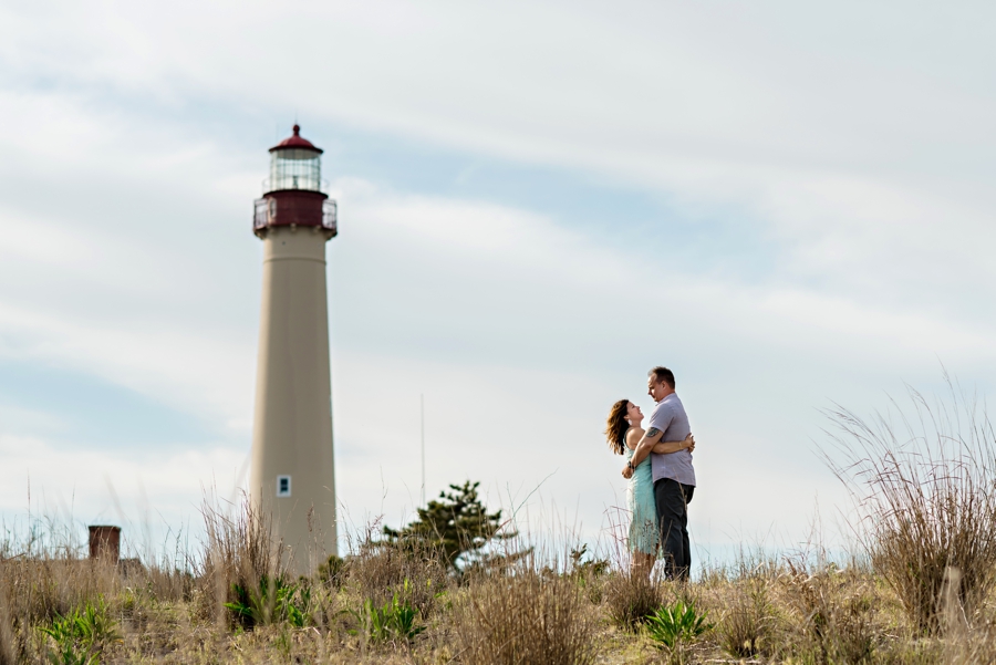 An engaged couple hold each other in front of a lighthouse on long beach island, nj.